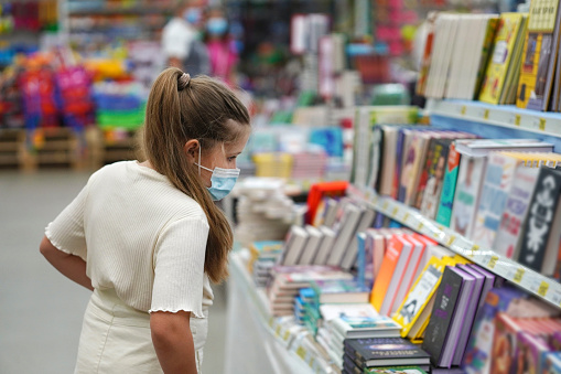 Happy smiling school girl in a protective medical mask chooses books in a bookstore. Prevention of coronavirus. Back to school education knowledge college university concept.