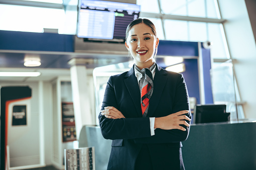 Portrait of a happy young woman working at airport. Flight attendant standing at boarding gate with her arms crossed at international airport.