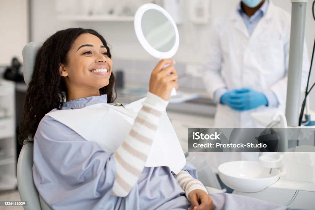 Happy Black Female Patient Looking At Mirror After Dental Treatment In Clinic Happy Black Female Patient Looking At Mirror After Dental Treatment In Clinic, Cheerful African American Woman Sitting In Chair In Stomatological Cabinet And Enjoying Her Beautiful Smile, Closeup Dentist Stock Photo