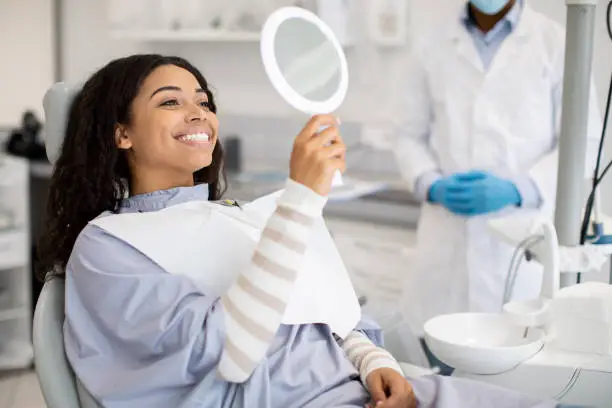 Happy Black Female Patient Looking At Mirror After Dental Treatment In Clinic, Cheerful African American Woman Sitting In Chair In Stomatological Cabinet And Enjoying Her Beautiful Smile, Closeup