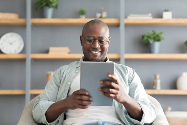 디지털 태블릿을 사용하여 행복한 흑인 성숙한 남자, 액세서리와 선반 위에 안락 의자에 앉아, 온라인 읽기 책 - front view happiness chair sitting 뉴스 사진 이미지