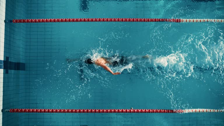 Aerial Top View Male Swimmer Swimming in Swimming Pool. Professional Athlete Training for the Championship, using Front Crawl, Freestyle Technique. Cinematic Wide Slow Motion Tracking Shot