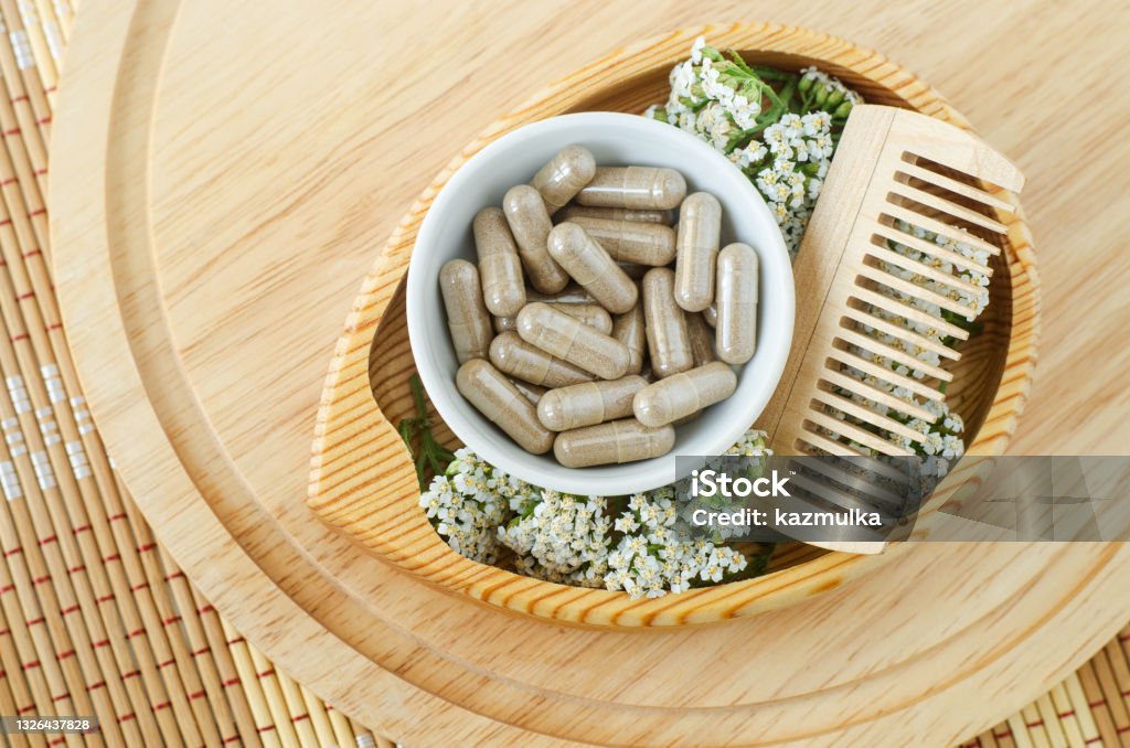 Small white bowl with food supplement capsules, wooden hairbrush and yarrow flowers. Natural healthcare, herbal medicine, haircare and beauty treatment. Top view, copy space. Hair Stock Photo