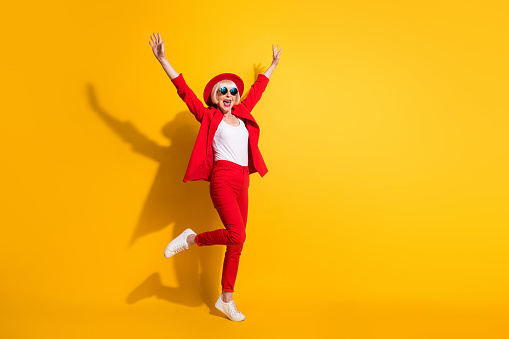 Full length body size photo of elder woman wearing stylish red suit sunglass smiling isolated on vibrant yellow color background.