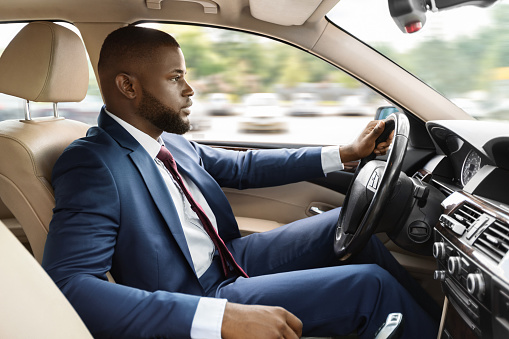 Handsome black businessman driving car, shot from going on business trip, side view, copy space. African american man in stylish suit going to business meeting, driving his luxury car