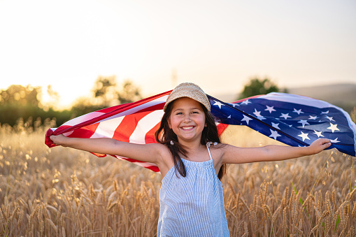 Proud and excited 6 year’s old girl, celebrating the 4th of July, while proudly holding the USA flag, during summer day, while standing in the wheat field