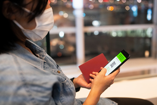 A female tourist is wearing a protective face mask and using a phone while waiting in an airport for her flight.