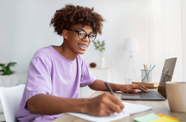 istock Home schooling concept. Smart black teen guy using laptop at table, writing in notebook during online lesson indoors 1326447543