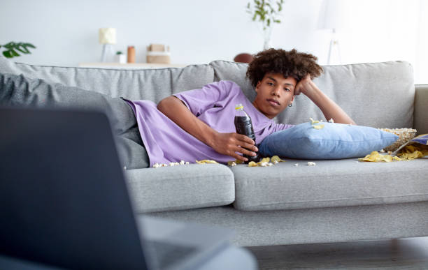 adolescente aburrido acostado en el sofá con bocadillos dispersos, viendo películas en la computadora portátil en casa - television boredom men sofa fotografías e imágenes de stock