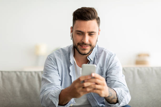 istock Young man with earbuds using smartphone, browsing internet and listening to music online, sitting on couch at home 1326450298