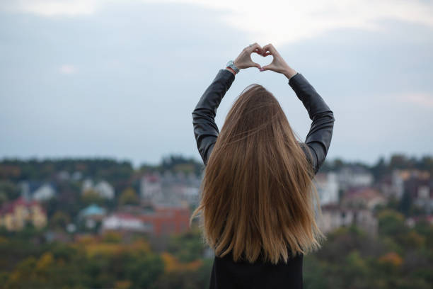 istock Pretty long haired model showing heart symbol with two hands over a city background. Space for text 1326450986