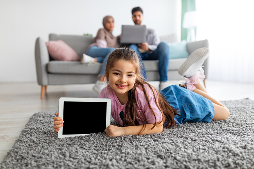 Little eastern girl showing digital tablet with empty screen, holding device with blank space for mockup, lying on floor carpet and smiling at camera. Parents relaxing on sofa with laptop