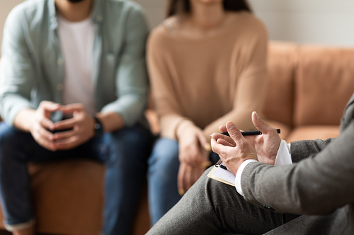 Help Concept. Closeup Cropped View Of Male Counselor Therapist Or Consultant In Suit Talking With Couple Sitting In Office, Giving Professional Advice. Selective Focus On Hands With Clipboard And Pen