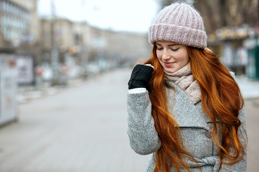 Closeup shot of amazing ginger woman with long hair wearing knitted cap and scarf walking at the city. Space for text
