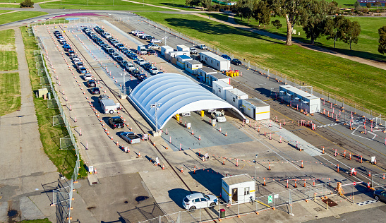 Pop-up Covid-19 testing centre in parklands on the edge of the city of Adelaide, South Australia: large canopy set on former racetrack with rows of people in cars, SUVs, taxis, waiting to receive a Covid-19 test.  Logos and ID edited.