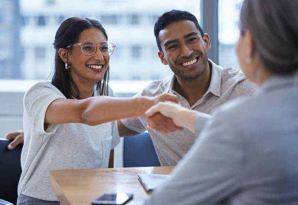 shot of a young couple sharing a handshake with a consultant they're meeting to discuss paperwork an office - bank stockfoto's en -beelden