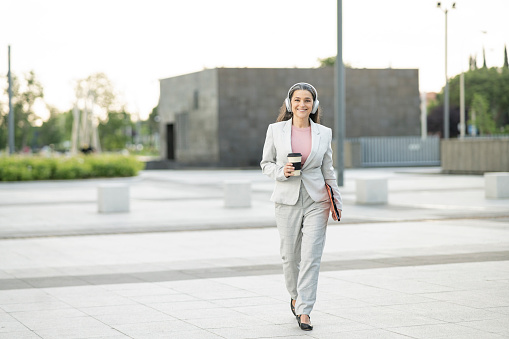 business woman walking through financial city with smartphone and coffee, with headphones