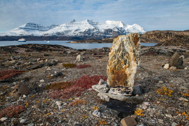 Photo of A large stone erected as a cairn, or orientations-marker, stands in a rocky area, with the expedition cruise ship MV Sea Spirit (Poseidon Expeditions) at anchor in front of snow-dusted mountains in the distance