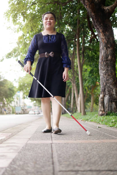 Asian blind person woman walking on sidewalk with a long white cane a mobility tool used to detect objects in the path, also helpful for onlookers in identifying the user as blind or vision disability mujer ciega asiática que camina por la acera con un bastón blanco largo, una herramienta de movilidad utilizada para detectar objetos en el camino, también útil para los espectadores en la identificación del usuario como ciego o discapacidad visual