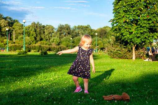 Cute little blond baby girl two year old playing with teddy bear on fresh green grass with flowers. Kid having fun making first steps on mowed natural lawn. Happy and healthy childhood concept