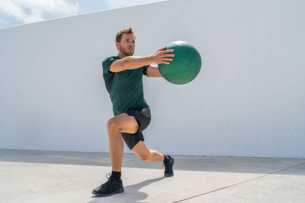 薬のボールの重量で突進ツイスト運動を行う男のトレーニング脚とコア腹筋トレーニングをワークアウト.腹筋トレーニングのために肺と胴体の回転をしているジムのアスリート。 ストックフォト