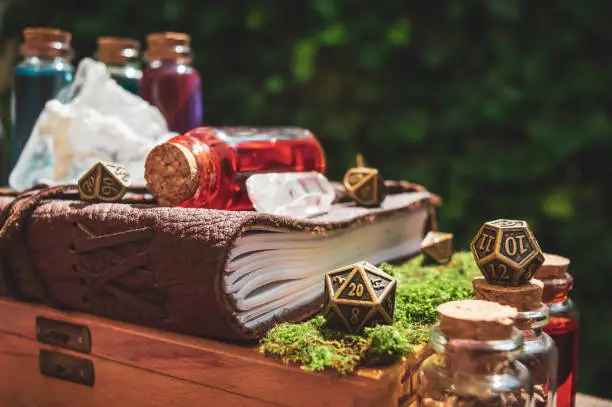 Close-up of a 20-sided metallic role-playing gaming die next to a leather-bound book, potion bottles, and crystals in the sun