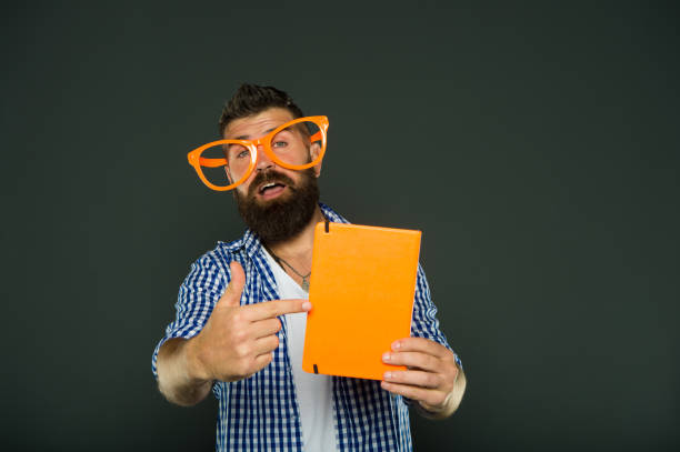 Look at this book. Bearded man in party glasses pointing at lesson book. Study nerd holding book. Book nerd wearing fancy glasses. University male student with lecture notes.