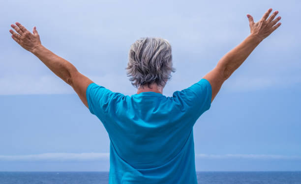 vue arrière de la femme âgée à bras ouverts devant la mer. horizon au-dessus de l’eau. ciel bleu - senior adult outdoors wellbeing sky photos et images de collection