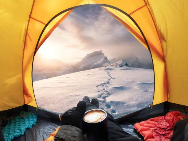 Photo of Man holding coffee cup and enjoying view of sunrise on snowy mountain inside yellow tent