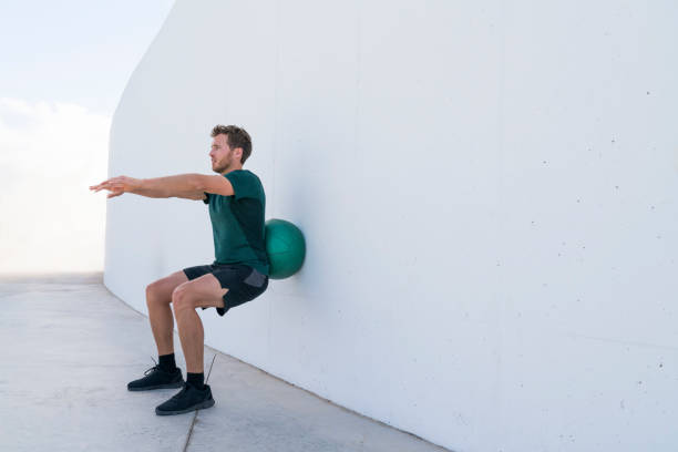 istock Strength training man doing squats using medicine ball rolling on wall squatting at fitness centre. Workout squat bodyweight exercises using medicine ball. Fitness athlete working out. 1326484880