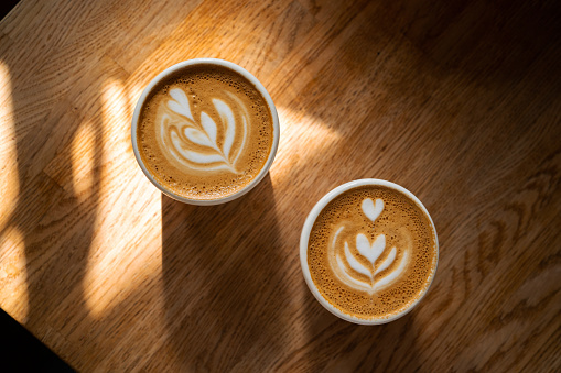 Two cups of fresh coffee with latte art on wooden table in sunlight. Morning cappuccino with hearts in cafe for couple. Coffee break concept. Caffeine drink for breakfast closeup. Top view, flat lay.