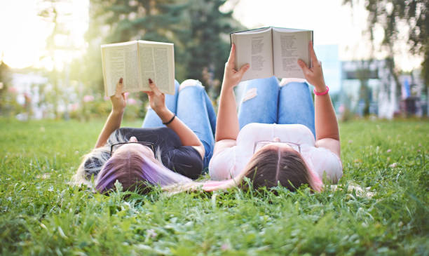 Teenagers reading books together in a university campus while lying in grass - Millennials learning and preparing to exam - Recreation and culture concept
