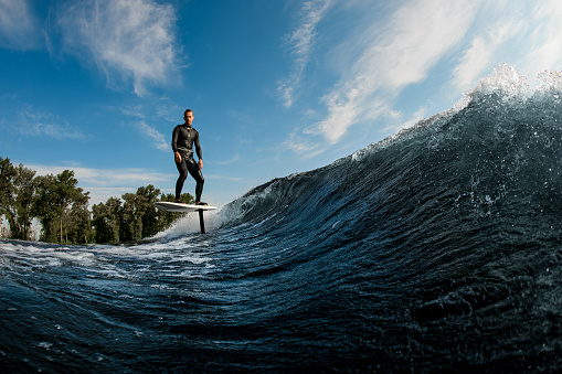 active man skillfully riding on the wave with hydrofoil foilboard on a background of blue sky and trees