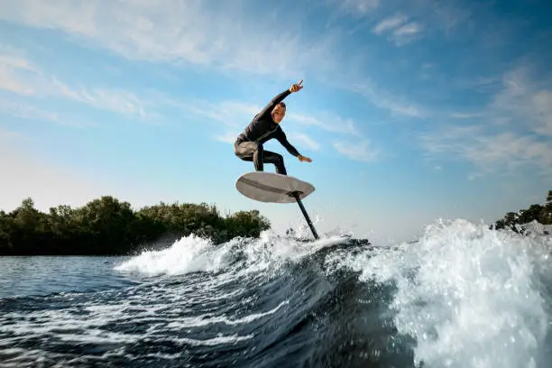young athletic man in black wetsuit balancing on a foilboard on a wave on a sunny day