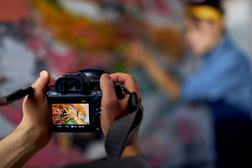 Close up of male hands holding professional digital camera on a blurred background. Female artist creating painting in the background. The concept of technology for working with creative photoshoot