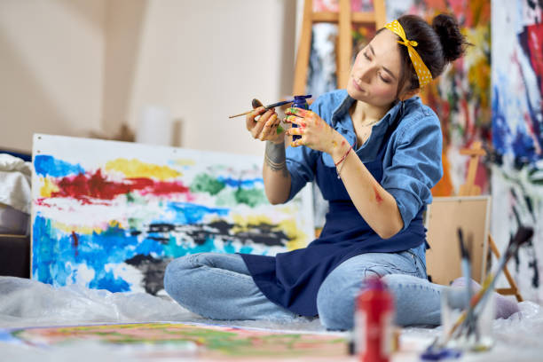 편안한 젊은 여성, 앞치마에 여성 화가 푸른 페인트 항아리와 페인트 브러시를 들고 추상화 작업하는 동안, 홈 워크샵에서 바닥에 앉아 - craft adult blue busy 뉴스 사진 이미지