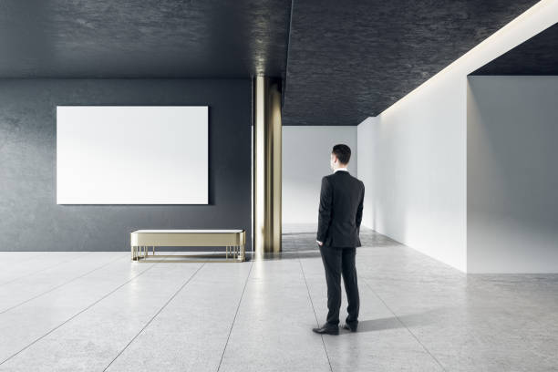 istock Businessman in modern gallery interior with empty poster on concrete wall. Mock up. 1326503533