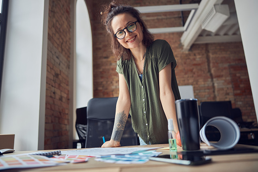 Creative woman, interior designer or architect in casual wear with messy hairdo smiling at camera while working on a blueprint for new project, standing in her office. Home decoration and renovation