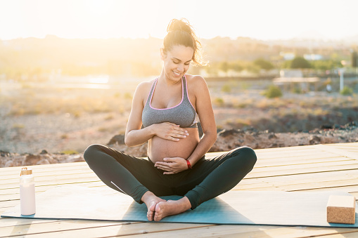 Young pregnant woman massaging the belly while doing yoga meditation outdoor - Health lifestyle and maternity concept