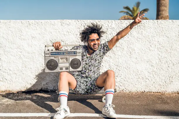 Young Afro Latin man having fun listening music with headphones and vintage boombox during summer vacations