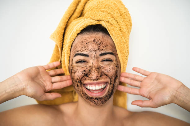 Young smiling woman applying coffee scrub mask on face - Happy girl having skin care spa day at home - Healthy alternative natural exfoliation treatment and youth people lifestyle concept stock photo