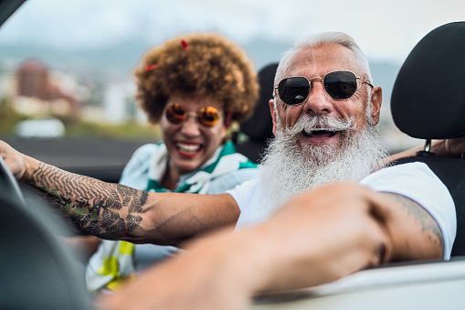 Happy senior couple having fun driving on new convertible car - Mature people enjoying time together during road trip tour vacation - Elderly lifestyle and travel culture concept