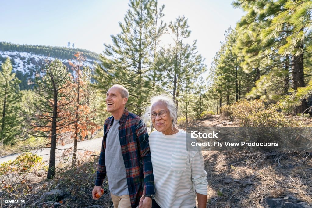 Active retired couple hiking in forest Wide angle shot featuring a Pacific Islander woman and her Caucasian husband enjoying nature on a sunny day. The active and healthy mixed race senior couple is hiking along a dirt trail through a forest. They are holding hands and smiling. Senior Adult Stock Photo