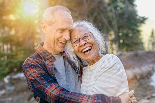 An active mixed race senior couple laugh while hiking together through a forest on a sunny day. They are on vacation and staying healthy while enjoying nature.