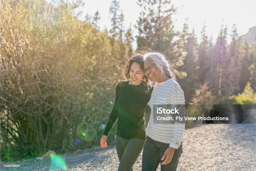 Portrait of beautiful mixed race senior woman spending time with her adult daughter outdoors A beautiful mixed race young adult woman embraces her vibrant retirement age mother. The mother and daughter are enjoying a relaxing walk in nature on a beautiful, sunny day. In the background is a mountainous evergreen forest bathed in sunlight. Senior Adult Stock Photo