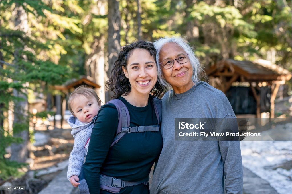 Happy multi-generation family enjoying nature hike A young mixed race woman carries her one year old daughter on her back in an ergonomic baby carrier while out on a hike with her senior mother. The multi-generation family is staying active and healthy outdoors in Oregon. Family Stock Photo
