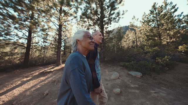 Wide angle tracking shot featuring a Pacific Islander woman and her Caucasian husband enjoying nature on a sunny day. The active and healthy mixed race senior couple is hiking along a dirt trail through a forest. They are holding hands and smiling.