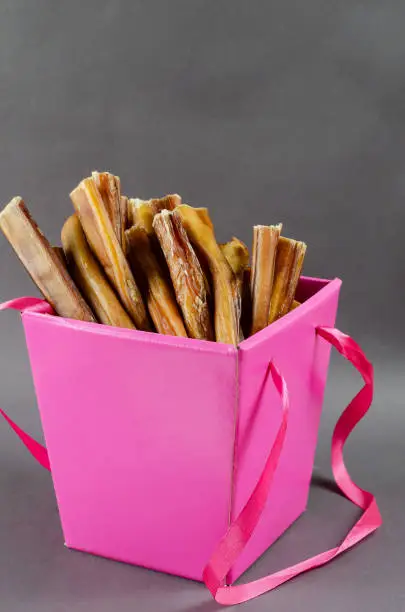 Photo of Beautiful pink gift box with pet treats. Open box full of bully sticks for dogs on gray background.