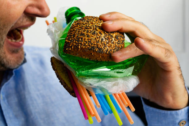 plastic in food A man eats a burger made from a tasty bun with sesame and poppyseed filled with different types of disposable plastic. The concept of disposable plastic as a danger for ecology and human health. microplastic stock pictures, royalty-free photos & images