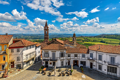 Small town square surrounded by houses with red roofs and belfries on background in Govone - commune overlooking green hills under blue sky with white clouds in Piedmont, Northern Italy.